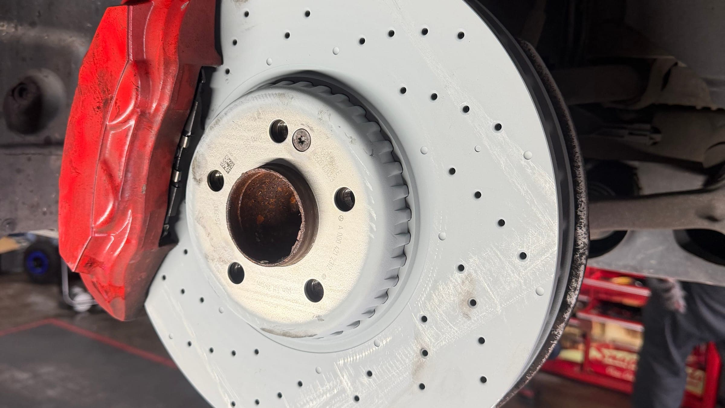 A cross-drilled Mercedes-AMG brake disc and red caliper with the wheel removed, the car raised on the workshop floor