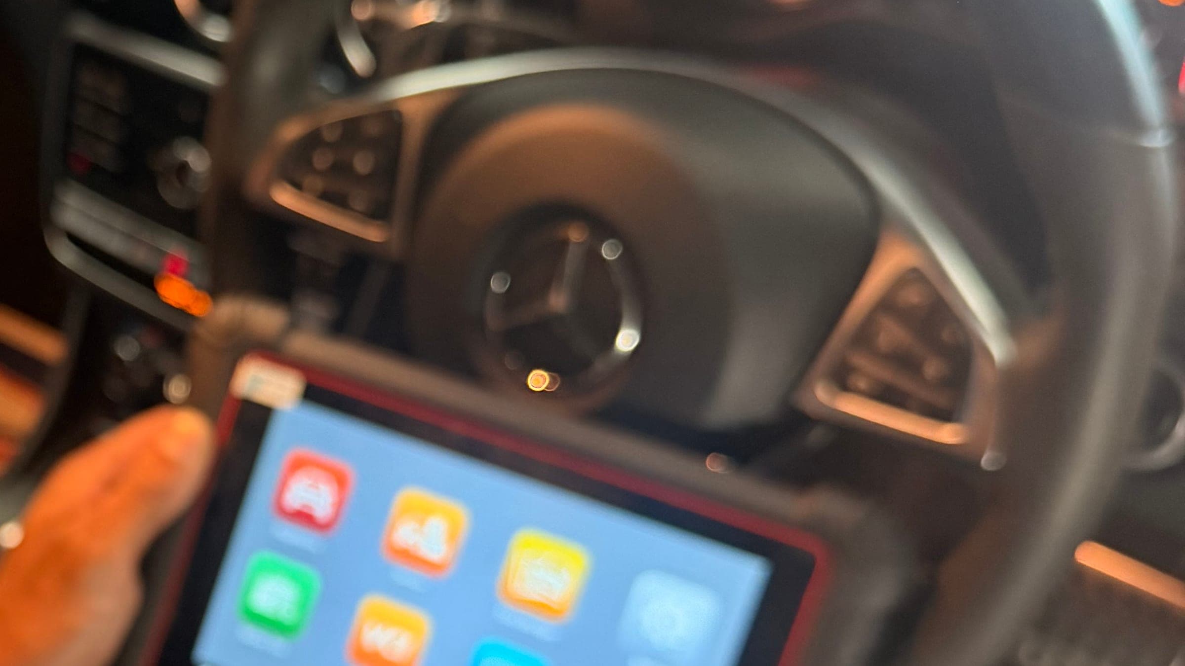 A diagnostic tablet plugged into a Mercedes and held in front of the steering wheel, with the instrument cluster lit