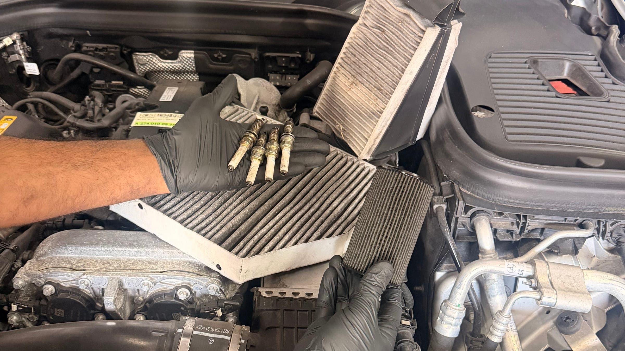 A technician in nitrile gloves holding a used cabin filter and an oil filter element over an open Mercedes engine bay, with four removed spark plugs laid out
